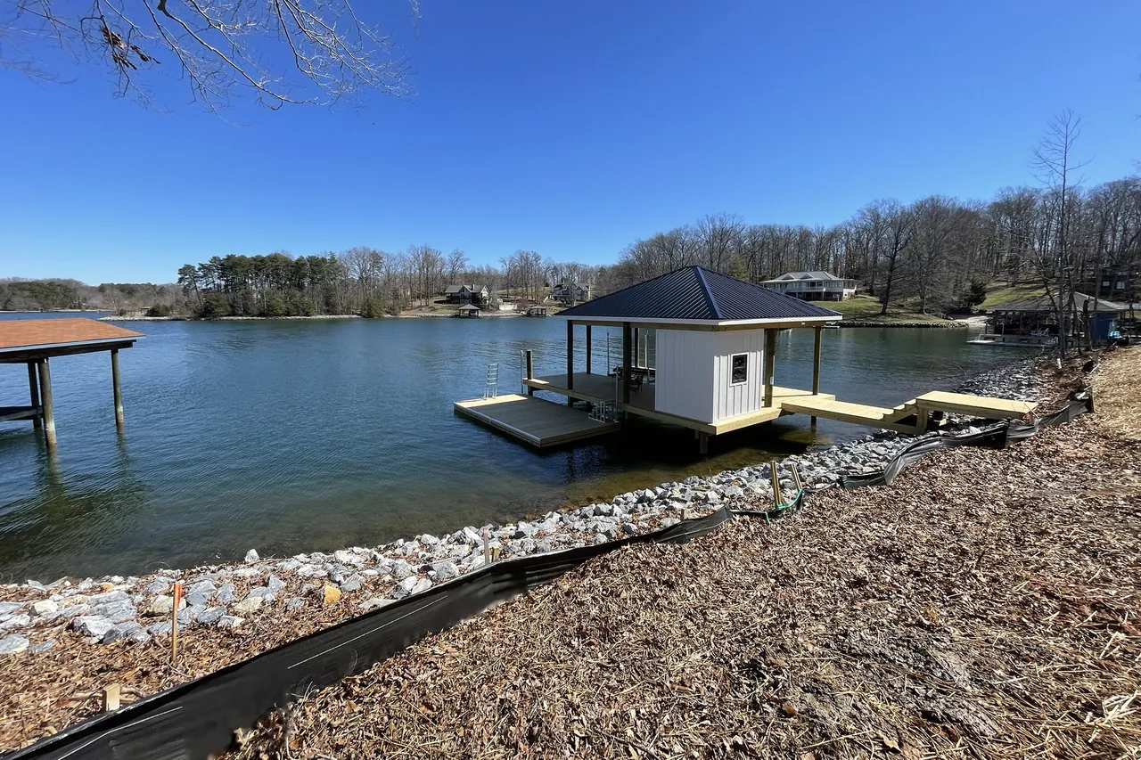 A boathouse with a dark metal roof and a floating dock, joined to the shore by a walkway and steps, above newly placed riprap and silt fence