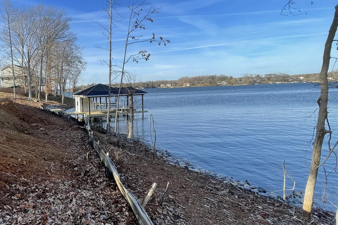 Graded bare ground sloping toward a lake, with a silt fence running between the soil and the shoreline and a boathouse beyond