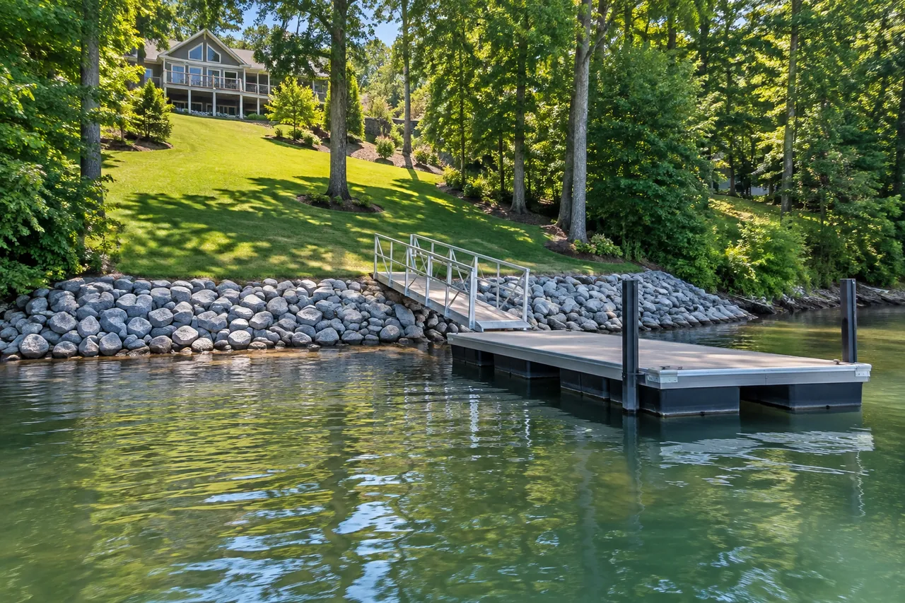 Sloped lawn running down to a band of gray riprap and a small floating dock with an aluminum ramp, below a lake home