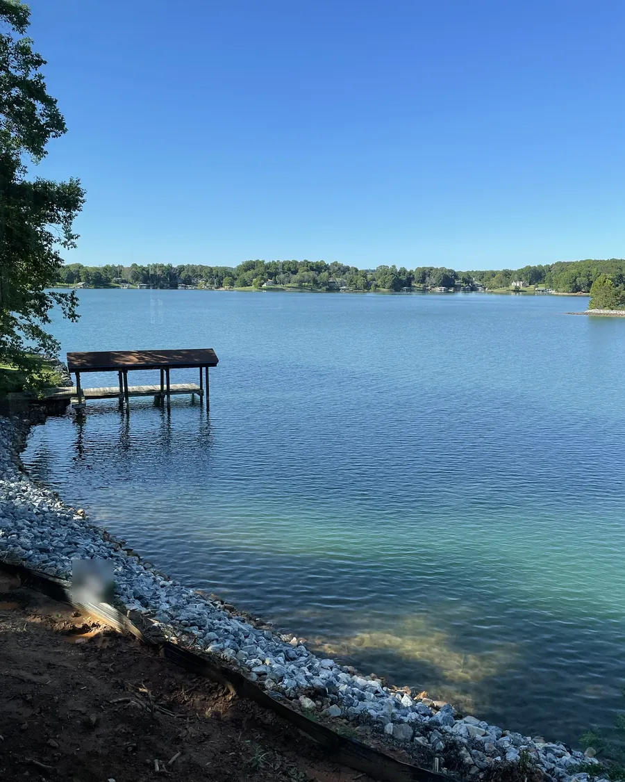 Newly placed riprap along a lake bank in summer, with silt fence on the bare soil above it and a covered dock past the end of the stone