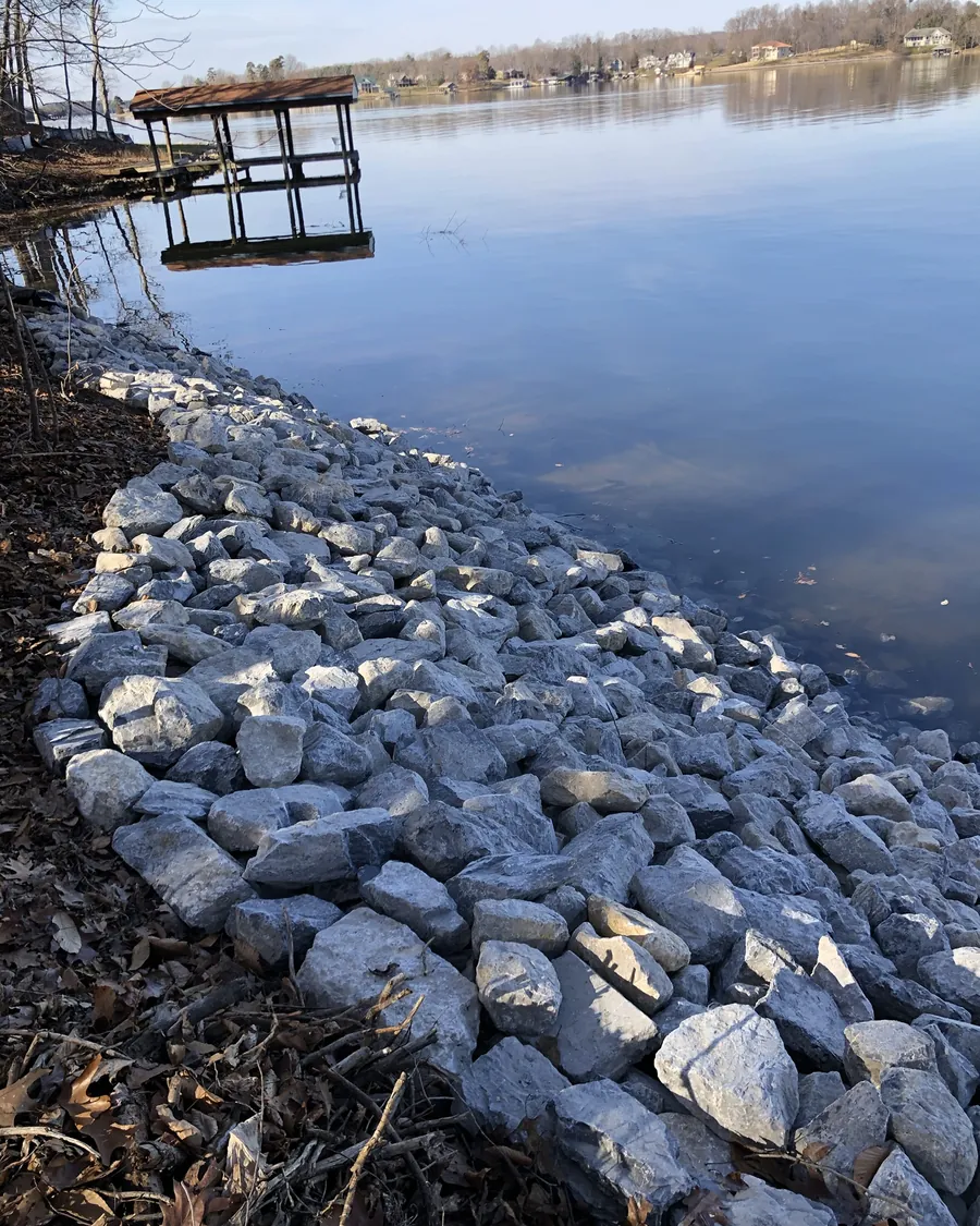 New gray riprap along a lake bank with a flagged stake at the top, and a covered dock out on the water