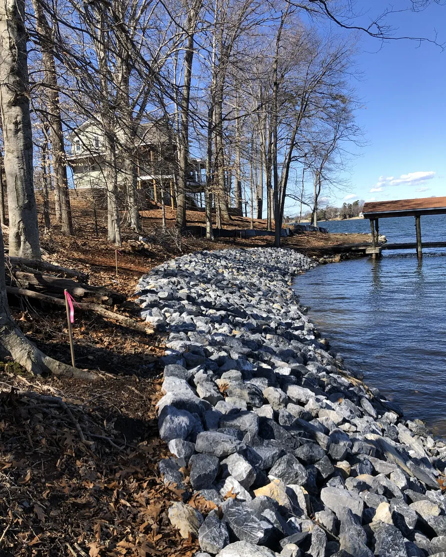 Gray riprap stone along a wooded lake bank below a house, with a covered pier past the end of the stone
