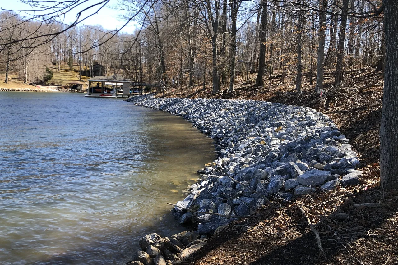 Riprap lining the shoreline of a narrow cove, with bare trees on the bank and boathouses at the far end