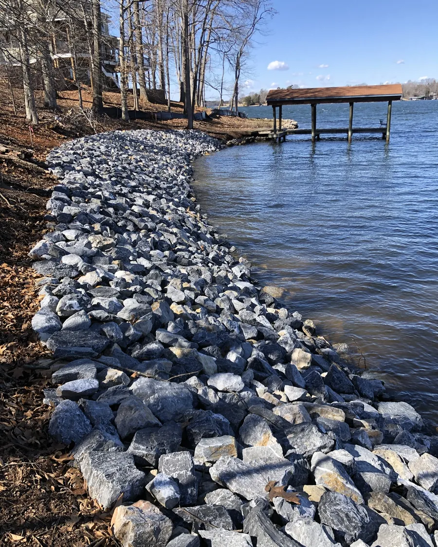 A long run of gray riprap stone along a lake bank under bare winter trees, leading out to a covered dock