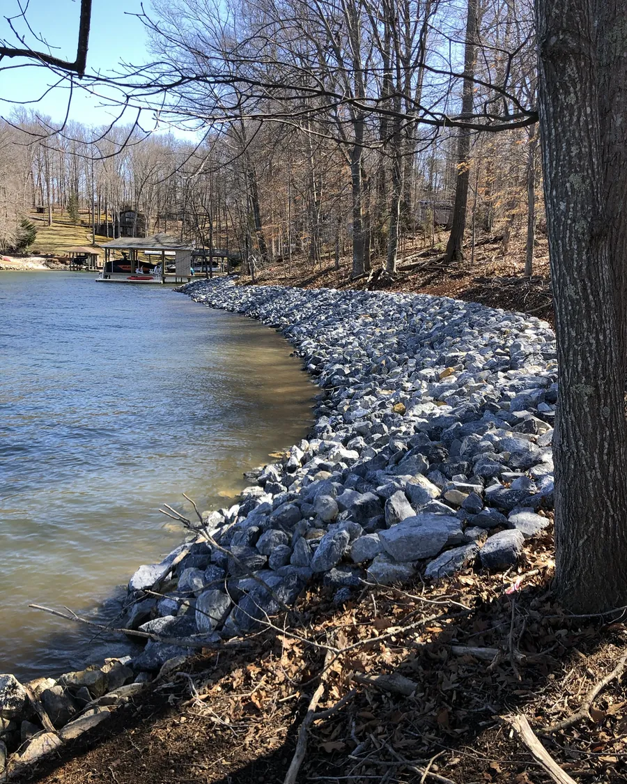 Riprap stone following the curve of a small lake cove, with bare winter trees above the bank