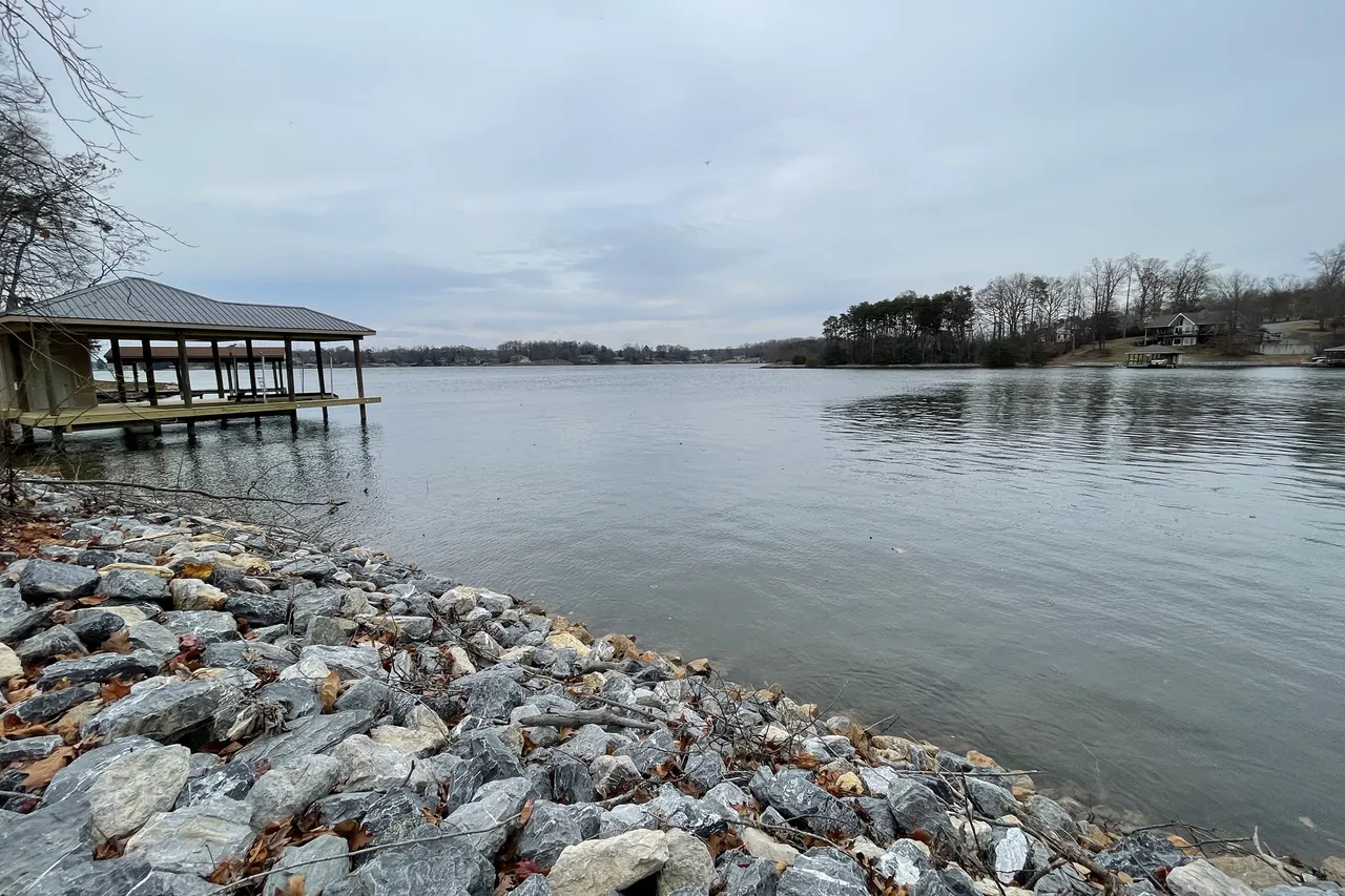 Riprap stone along the edge of the lake on a gray winter day, with a covered boathouse on the shore and the open lake beyond