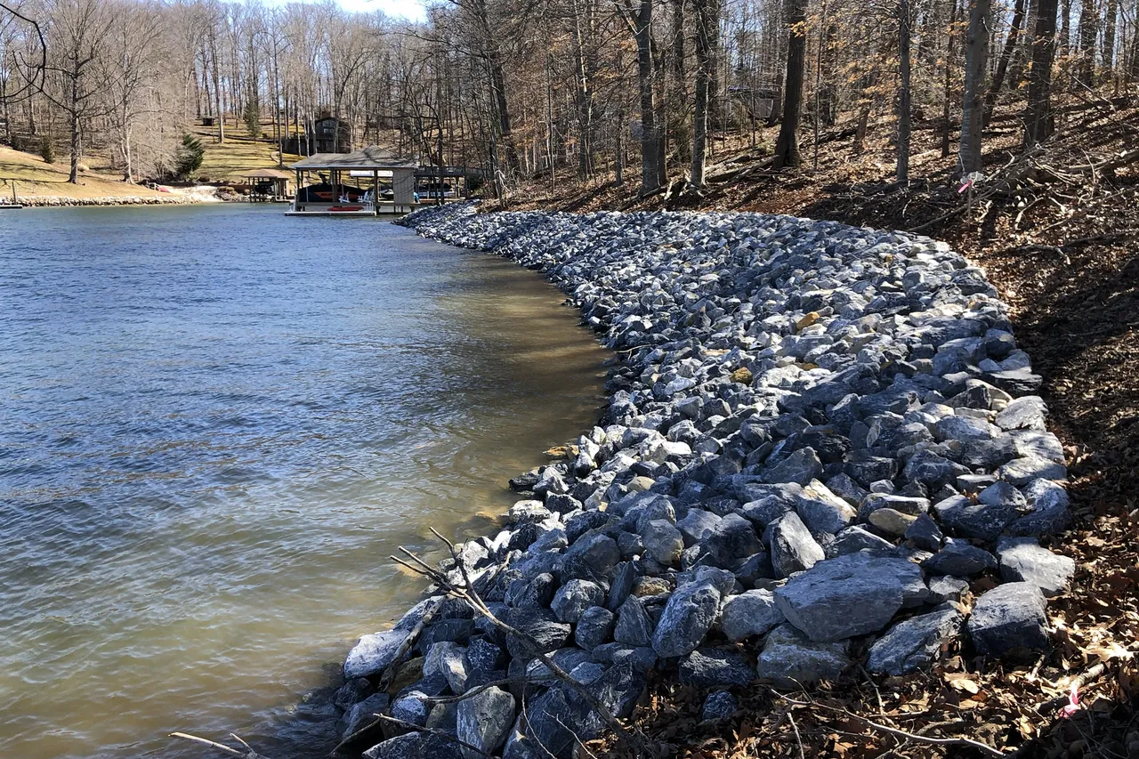 New gray riprap following the curve of a wooded cove, with the trees above it left standing and a covered dock at the end of the cove